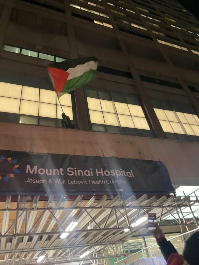Photo of person on top of the entrance to Mount Sinai Hospital in Toronto at night, having a Palestinian flag.
